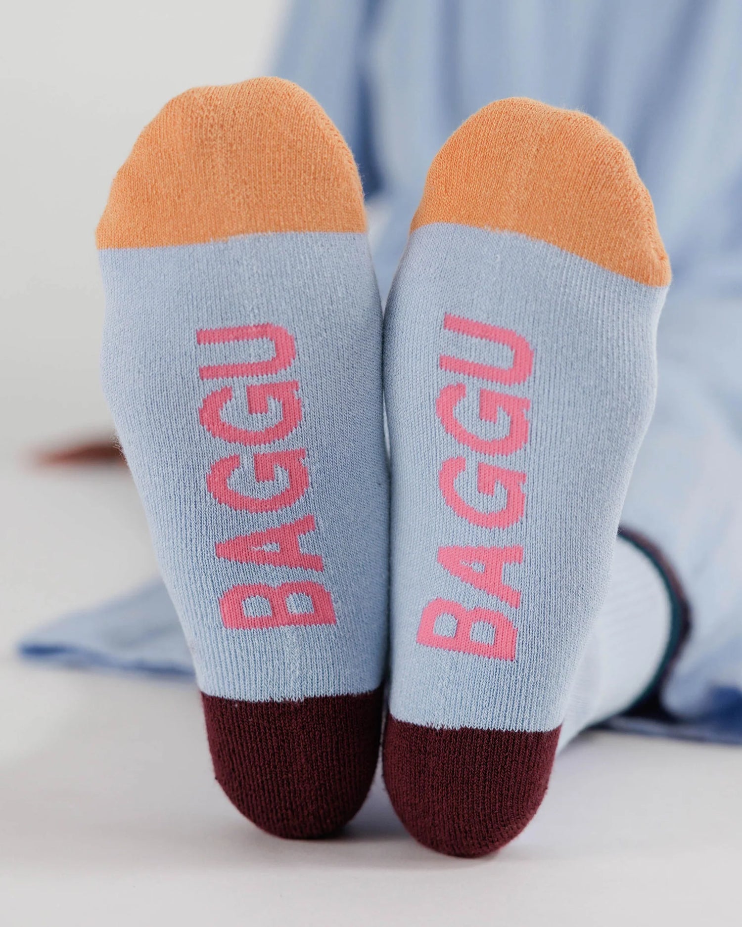 Light Blue Socks with black Ankle Stripe, Brown heel, orange colour blocked toes, and pink "BAGGU" text on bottom of feet